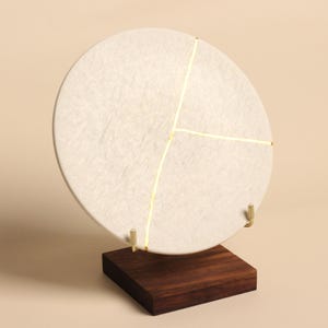 May include: A circular off-white textured disc featuring a gold-colored vein pattern that divides the surface into three sections. The disc is supported by two small gold-colored metal hooks attached to a square dark brown wooden base stand.