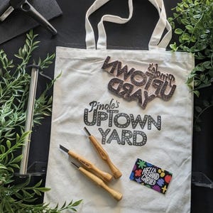 May include: Cream-colored tote bag with the text "Pinole's Uptown Yard" and a wooden stamp. Three wood-handled carving tools and a colorful sticker are also visible. A black roller and greenery are in the background.