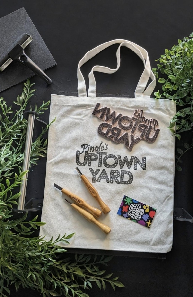May include: Cream-colored tote bag with the text "Pinole's Uptown Yard" and a wooden stamp. Three wood-handled carving tools and a colorful sticker are also visible. A black roller and greenery are in the background.