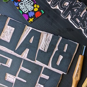 May include: A close-up shot of a black block with the word "HELLA" carved out in white. A business card with the text "HELLA HAND CARVED" and colorful shapes is visible. Carving tools and a carved block with the word "BLACK" are also present.
