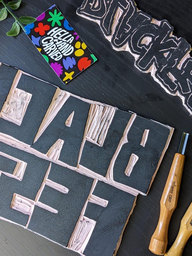 May include: A close-up shot of a black block with the word "HELLA" carved out in white. A business card with the text "HELLA HAND CARVED" and colorful shapes is visible. Carving tools and a carved block with the word "BLACK" are also present.