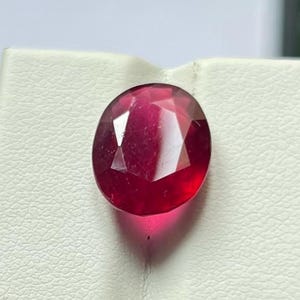 Puede incluir: Una gema de rubí facetada de forma ovalada. La gema es de un color rojo intenso y rico y se muestra sobre una superficie blanca. El corte del rubí crea múltiples facetas que reflejan la luz, dándole una apariencia brillante.