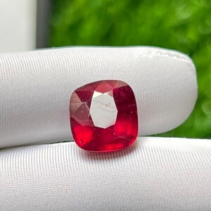 May include: A cushion-cut, faceted, red gemstone. The gem is held in a white gloved hand, with a blurred green background. The gemstone has a bright, reflective surface.