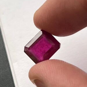 May include: A faceted, rectangular-cut ruby gemstone. The gem is held between two fingers, showcasing its deep magenta color and sharp edges. The ruby's facets reflect light, highlighting its clarity and brilliance. The background is a neutral white and gray.