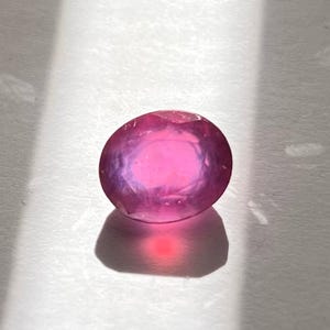 Op de afbeelding: Een gefacetteerde, ovaalvormige edelsteen met een levendige roze tint. De edelsteen ligt op een witte ondergrond, met schaduwen die over de achtergrond vallen. De facetten van de steen vangen het licht op en creëren een sprankelend effect.