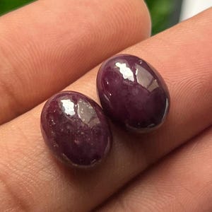 Puede incluir: Dos piedras preciosas cabujón de forma ovalada, de color púrpura intenso, con una superficie lisa y pulida. Cada piedra presenta un brillo reflectante y un aspecto sólido y opaco, mostrando un tono violeta oscuro constante en las formas redondeadas.