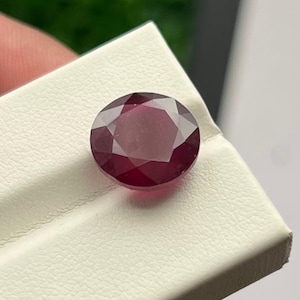 May include: A faceted, round, deep red gemstone, likely a ruby, rests on a white surface. The gem's cut reveals multiple facets, enhancing its brilliance. The background is blurred, focusing attention on the vibrant jewel.