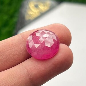 Op de afbeelding: Een ronde, gefacetteerde, felroze edelsteen. De edelsteen wordt in een hand gehouden, waarbij de briljante slijpvorm en levendige kleur worden getoond. Het oppervlak van de edelsteen reflecteert licht, waardoor de vele facetten worden benadrukt. De achtergrond is wazig.