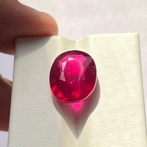 Könnte beinhalten: Ein lebendiger, oval geschliffener Rubin-Edelstein. Der Edelstein hat eine tiefe, satte rote Farbe mit Facetten, die das Licht einfangen. Der Rubin ist vor einem weißen Hintergrund platziert, der seine Brillanz zeigt. Dieser Edelstein ist ein schönes Beispiel für feinen Schmuck.