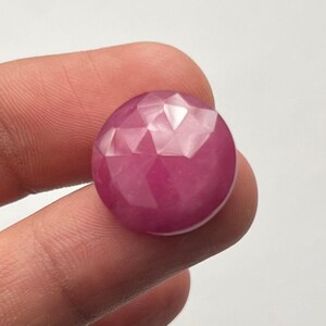 Op de afbeelding: Een gefacetteerde, ronde robijn edelsteen. De edelsteen heeft een dieproze-rode kleur en een gepolijst, reflecterend oppervlak. De facetten creëren een geometrisch patroon over het oppervlak van de steen. De edelsteen wordt tussen twee vingers gehouden.