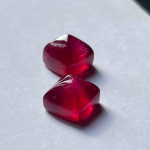 May include: Two faceted, deep red gemstones with a square-like shape. The gems are highly polished, reflecting light and appearing translucent. They are set against a white background, highlighting their rich colour and clarity. Ideal for jewellery making.