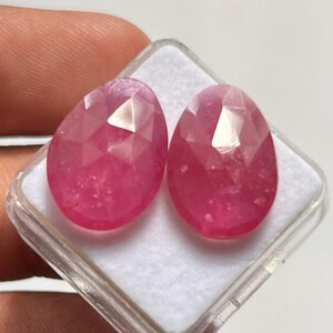 Könnte beinhalten: Zwei ovale, rosa Edelsteine mit Facettenschliff, präsentiert in einer transparenten Kunststoffbox. Die Edelsteine haben einen Brillantschliff, der das Licht reflektiert. Die Box liegt auf einer weißen Oberfläche.