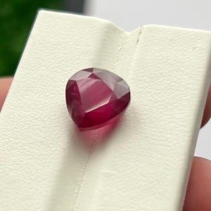 May include: A faceted, heart-shaped ruby gemstone. The ruby is a deep, rich red color and is displayed on a white surface. The gemstone's facets catch the light, creating a sparkling effect. The ruby is a precious stone.