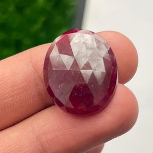 Puede incluir: Una gema de rubí facetada de forma ovalada. La gema es de color rojo intenso con múltiples facetas que reflejan la luz. El rubí se sostiene en una mano, mostrando su tamaño y forma. El fondo está borroso, centrando la atención en la gema.