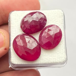 Puede incluir: Tres gemas facetadas, de forma ovalada y de color rojo intenso, se muestran en una caja de plástico transparente. Las gemas tienen un corte brillante, que refleja la luz. La caja está sobre una superficie blanca.