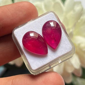 Op de afbeelding: Twee traanvormige, levendig magentarode edelstenen met een glad, gepolijst oppervlak. De stenen worden naast elkaar gepresenteerd in een kleine, vierkante, doorzichtige plastic opbergdoos met een witte schuimrubberen inzet voor veilige plaatsing.