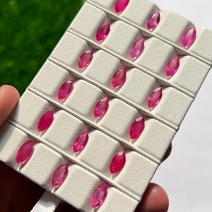 Könnte beinhalten: Eine Sammlung von sechzehn losen, marquise-geschliffenen rosa Edelsteinen, die in einem weißen Präsentationstablett mit einzelnen Fächern angeordnet sind. Jeder Edelstein hat eine facettierte Oberfläche, die das Licht reflektiert.
