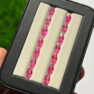 Puede incluir: Una colección de catorce piedras preciosas de color rosa vibrante, cada una tallada en forma de marquesa con superficies facetadas. Las piedras están dispuestas en dos filas verticales paralelas dentro de un estuche negro con un inserto blanco texturizado.