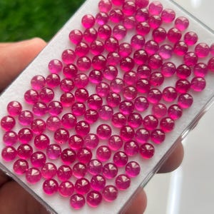 Peut inclure: Une collection de nombreuses petites pierres précieuses cabochons, rondes et d'un rose vif, disposées en grille sur un plateau de présentation en mousse blanche. Chaque pierre présente une surface bombée, lisse et polie qui reflète la lumière.