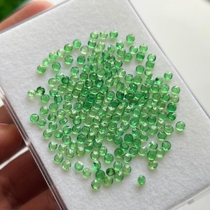 May include: A collection of numerous small, round, faceted green gemstones arranged in a cluster on a white foam insert inside a clear plastic display case. Each individual stone exhibits a brilliant, translucent emerald green hue.