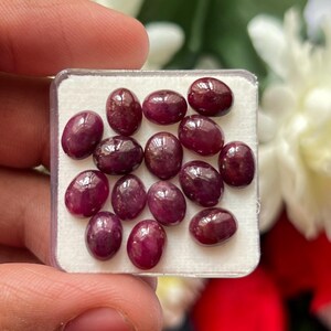 May include: A collection of thirteen oval-shaped, deep red rubies arranged on a white foam insert within a clear plastic display case. Each gemstone features a smooth, polished surface with a reflective luster, showcasing a consistent dark crimson hue.