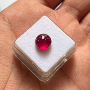 Könnte beinhalten: Ein kleiner, runder Edelstein im Cabochon-Schliff mit einer leuchtenden, tiefroten Farbe. Der Edelstein liegt auf einer weißen Schaumstoffeinlage in einer quadratischen, durchsichtigen Kunststoff-Präsentationsbox und zeigt seine glatte Oberfläche.