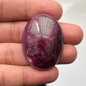 Könnte beinhalten: Ein ovaler Cabochon-Edelstein mit einer tief magentafarbenen und dunkelroten Farbpalette, sichtbaren schwarzen Einschlüssen und einer glatten, polierten Oberfläche, die das Licht auf seiner abgerundeten, glänzenden Oberseite reflektiert.