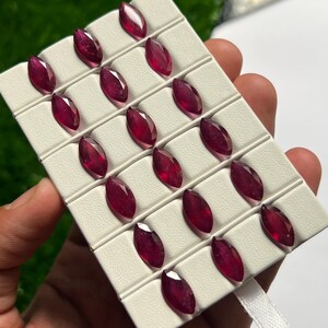 Könnte beinhalten: Eine Ansammlung von fünfzehn tiefroten, im Marquise-Schliff facettierten Edelsteinen, die in einem Raster auf einem weißen Display-Tablett angeordnet sind. Jeder Stein hat eine facettierte Oberfläche, die das Licht reflektiert.