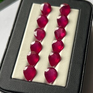 Può includere: Una collezione di dodici rubini sfaccettati a forma di pera, disposti in due file verticali da sei. Ogni gemma presenta un colore rosso intenso e vibrante con una superficie lucida e riflettente, presentata all'interno di un cofanetto nero.