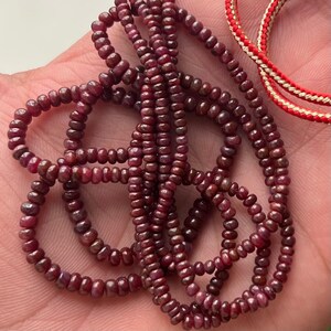 Pode incluir: Close-up de fios de contas de rubi vermelho escuro. As pequenas contas redondas são enfiadas, possivelmente para um colar ou pulseira. A cor é um vermelho escuro rico com um ligeiro brilho. Um cordão vermelho e dourado é visível no fundo.