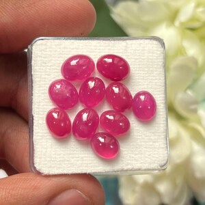Könnte beinhalten: Eine Ansammlung von neun ovalen, leuchtend pink-roten Cabochon-Edelsteinen, die auf einer weißen, strukturierten Schaumstoffeinlage in einer quadratischen, durchsichtigen Kunststoff-Präsentationsbox angeordnet sind.