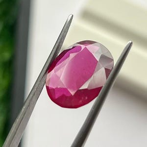 Könnte beinhalten: Ein facettierter, ovalförmiger Rubin-Edelstein, der von einer Pinzette gehalten wird. Der Rubin zeigt einen tiefen rosaroten Farbton mit helleren rosa und weißen Facetten. Der Edelstein ist der Mittelpunkt, die Pinzette sorgt für Kontrast.