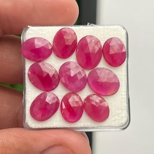 May include: A collection of nine oval-shaped, faceted pink rubies arranged in a grid pattern inside a clear plastic display case with a white foam insert. Each gemstone features a polished surface with multiple triangular facets reflecting light.