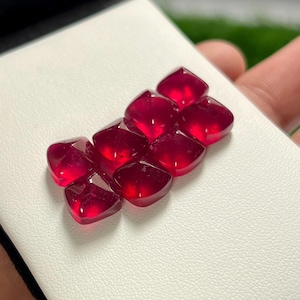 May include: A collection of seven loose, cushion-cut rubies with a deep, vibrant red color and faceted surfaces. These gemstones are arranged in a cluster, showcasing their translucent quality and polished finish, reflecting light across their edges.