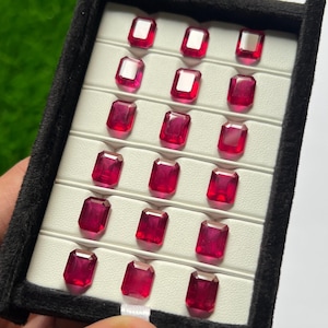 Puede incluir: Una colección de diecisiete rubíes rojos rectangulares y facetados dispuestos en un estuche negro con un inserto blanco. Cada piedra preciosa presenta una superficie pulida y un color rojo intenso, organizada en filas dentro de ranuras individuales.