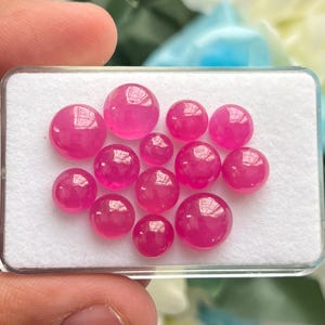 May include: A collection of twelve round, cabochon-cut gemstones in a vibrant, translucent hot pink color. The stones are arranged in a cluster on a white foam insert inside a clear rectangular display case, showcasing smooth, polished surfaces.