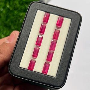 Può includere: Una custodia espositiva rettangolare nera contenente dieci pietre preziose sfaccettate, con taglio a baguette e di un vibrante colore rosso rosato, disposte in due file verticali parallele da cinque su un inserto bianco.