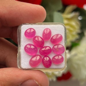 May include: A collection of nine oval-shaped, smooth, vibrant pink cabochon gemstones arranged in a square plastic display case with a white foam insert. Each stone features a polished, reflective surface with a consistent deep pink hue.
