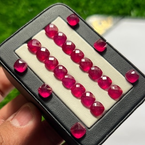 May include: A collection of small, faceted, cushion-cut vibrant red gemstones arranged in two parallel rows on a white insert within a black display box. The gemstones exhibit a brilliant, translucent quality with multiple facets reflecting light.