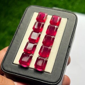 Pode incluir: Uma coleção de dez pedras preciosas vermelho-vibrantes, com corte esmeralda, dispostas em duas filas paralelas de cinco dentro de um estojo retangular preto com um inserto branco aveludado para segurar cada pedra no lugar.