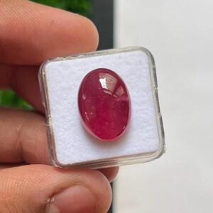Könnte beinhalten: Ein polierter, oval geformter Rubin-Edelstein mit tiefroter Farbe und einer glatten Oberfläche im Cabochon-Stil. Der Edelstein befindet sich sicher in der Mitte einer kleinen, quadratischen, durchsichtigen Kunststoff-Präsentationsbox mit weißem Schaumstoffeinsatz.