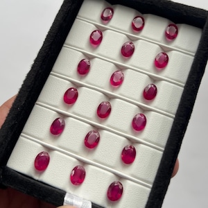 Peut inclure: Une collection de dix-sept rubis rouge foncé taillés en ovale, disposés dans un présentoir noir à intérieur blanc. Chaque pierre précieuse présente une surface à facettes qui reflète la lumière, révélant une teinte cramoisie vive et uniforme.