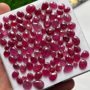May include: A collection of numerous small, polished, oval and pear-shaped rubies in varying shades of deep red and magenta. The gemstones are arranged closely together on a white display board, showcasing their smooth, reflective surfaces.