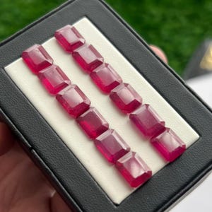 Può includere: Una collezione di gemme rosso scuro, tagliate a forma rettangolare, esposte in una custodia nera con interno bianco. Le gemme sono disposte in due file parallele, mostrando le loro superfici lucide e il loro colore vibrante. La custodia è leggermente inclinata.