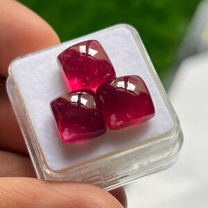 Può includere: Tre pietre preziose rosso intenso, tagliate a cuscino con una superficie liscia e lucida. Le pietre sono disposte in formazione triangolare all'interno di una piccola scatola espositiva quadrata in plastica trasparente con inserto in schiuma.