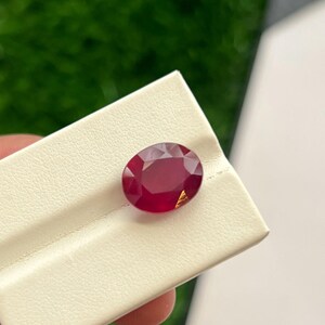 Peut inclure: Une pierre précieuse rouge profond de taille ovale avec une surface à facettes, posée sur un présentoir blanc. La pierre présente une petite inclusion ou un reflet de couleur dorée près de son bord inférieur, contrastant avec la teinte cramoisie.