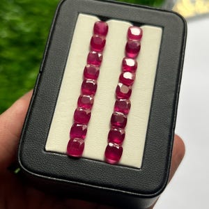 May include: A black rectangular display box containing two vertical rows of cushion-cut, deep pinkish-red gemstones. Each row consists of nine faceted stones arranged neatly on a white insert, showcasing their reflective surfaces and vibrant color.