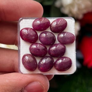 May include: A collection of nine oval-shaped, deep magenta-red cabochon gemstones arranged in a square clear plastic display case with a white foam insert. Each stone features a smooth, polished, domed surface with subtle natural inclusions.
