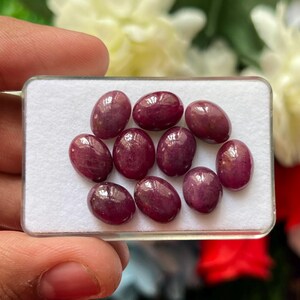 Puede incluir: Un conjunto de diez piedras preciosas cabujón de forma ovalada y color rojo intenso, dispuestas sobre una base de espuma blanca dentro de una caja de presentación de plástico transparente. Cada piedra tiene una superficie pulida y lisa.