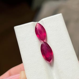 以下が含まれることがあります： 鮮やかな深いピンクレッドのマーキスカットが施された2つの宝石。これらのファセットカットされた石は、尖った端と対称的な細長い楕円形のシルエットが特徴で、表面に内部からの光の反射を伴う半透明の質感を備えています。